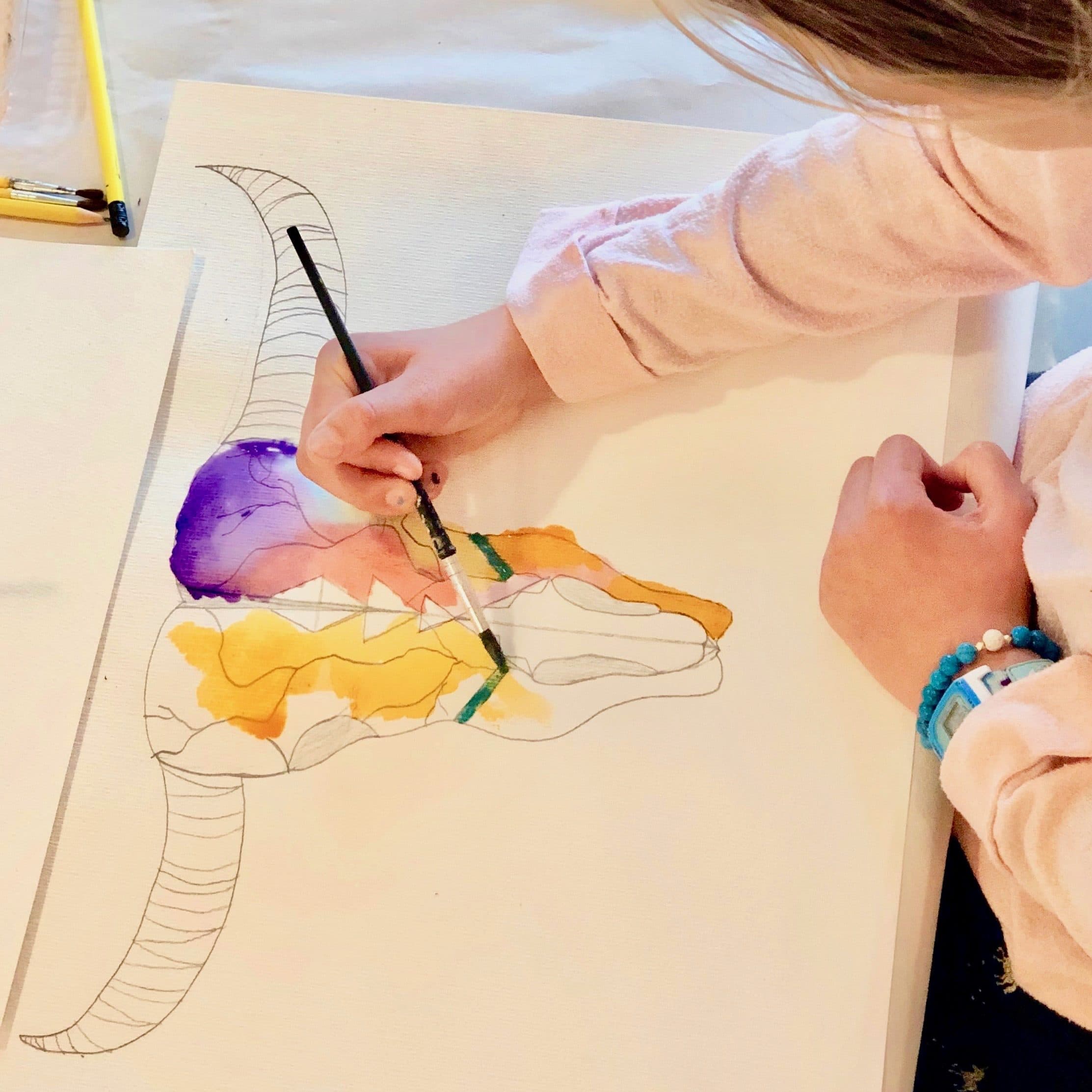A student paints a watercolor wash in purple and gold inside a line drawing of a skull